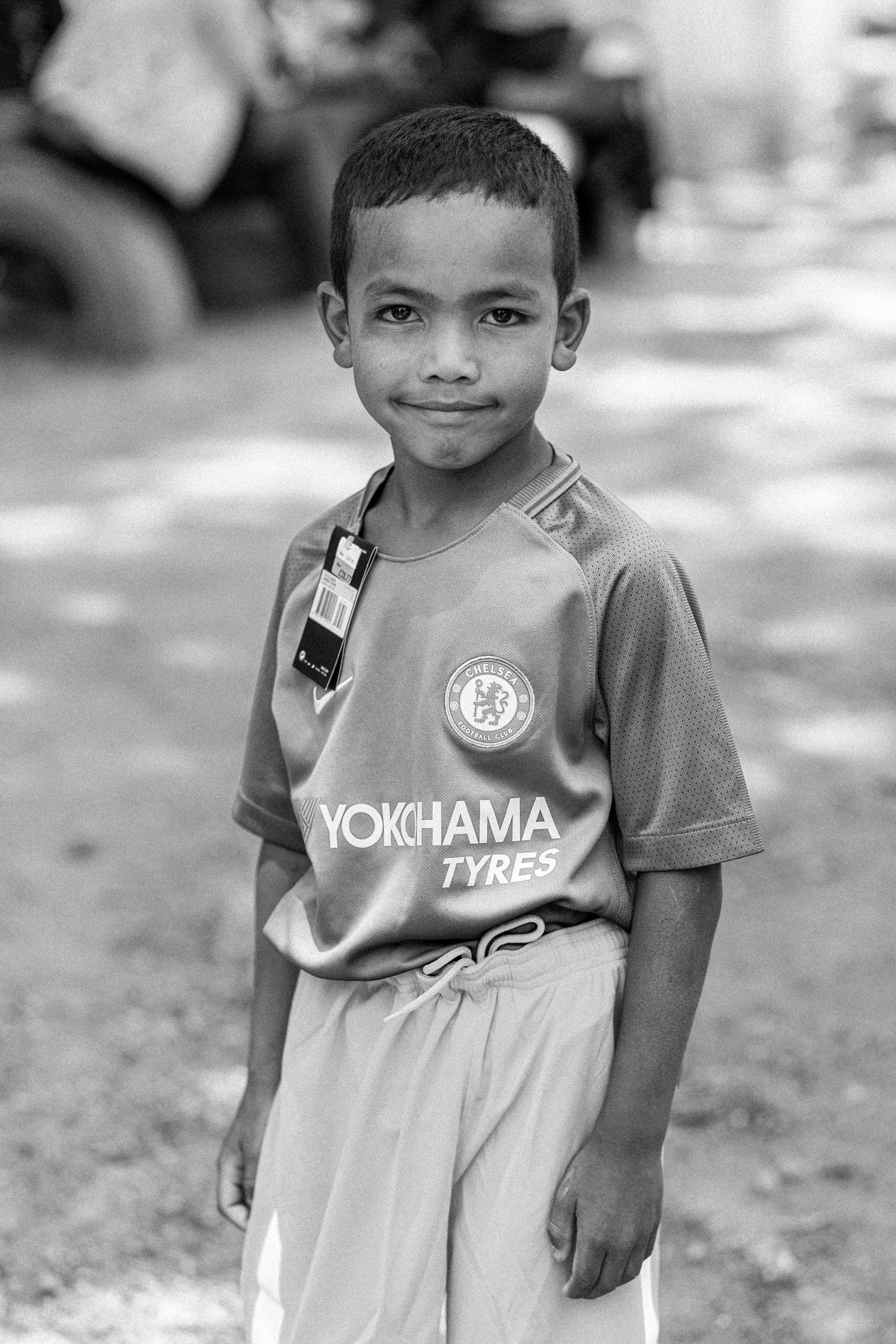 A black-and-white photo of boy wearing a Chelsea football shirt, smiling at the camera.